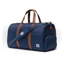 Load image into Gallery viewer, A navy blue Herschel duffle bag with brown handles and a logo on a white background. The adjustable shoulder strap is laid across the front.