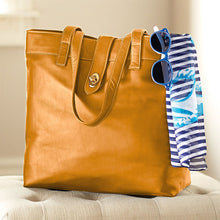 Load image into Gallery viewer, Angled front image of the tan leather tote bag. A pair of sun glasses and a blue striped scarf are hanging from the corner.