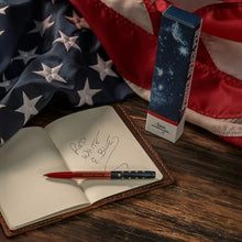 Load image into Gallery viewer, The pen displayed on top of a notebook that reads red white & blue on top of a wooden surface and an American Flag