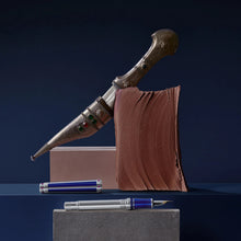 Load image into Gallery viewer, The Fountain Pen is placed without the cap and with its barrel off, showing the ink piston barrel. On the blue background, a soft light coats some Ottoman-themed decorations.