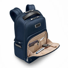 Load image into Gallery viewer, Angled view of the navy blue backpack. The bottom front pocket is unzipped. A passport, pen, USB cable, notebook, and keys on a keys sling are organized in different compartments.