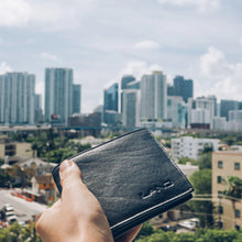 Load image into Gallery viewer, A hand holding the black leather wallet with a blurry urban city on the background.