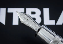 Load image into Gallery viewer, A close-up of the fountain pen nib and upper barrel against a black background with large white blurred letters.
