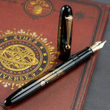 Load image into Gallery viewer, Angled view of the Fountain Pen. The cap is set off and standing on the left side. It is placed on top of a red book cover featuring an elaborate decoration with an Astrology design. It reads: "THE UNIVERSE"