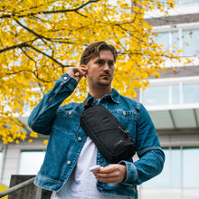 Load image into Gallery viewer, Man wearing a denim jacket with a black crossbody bag in an urban setting with yellow-leaved trees.