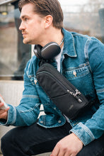 Load image into Gallery viewer, Man wearing a black crossbody bag and headphones, sitting outdoors.
