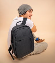 Load image into Gallery viewer, A young man sitting on a beige background while holding the black backpack.