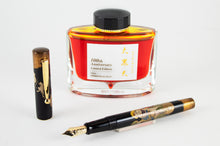 Load image into Gallery viewer, The Daikokuten Fountain Pen arranged with the yellow-orange ink bottle behind it.