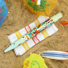 Load image into Gallery viewer, The image shows the new Retro 51 Beachy Side Up Rollerball Pen laying on a beach theme set with a small towerl, some umbrellas, and a small surf board.