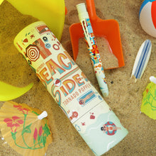 Load image into Gallery viewer, The Beachy Side Up Pen with a small surfboard, a bucket and pail, a beach ball, and the gift tube, arranged on a sandy background.