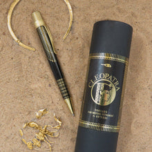 Load image into Gallery viewer, Side view of the rollerball pen on a sandy background with some golden jewelry, and the gift tube.