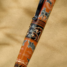 Load image into Gallery viewer, A close-up of the pen with an orange body, black bands, and illustrated motifs including snakes, bird, and warrior, resting on a textured beige surface.