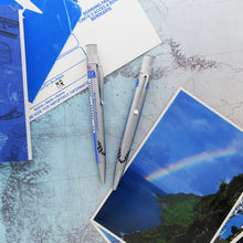 Load image into Gallery viewer, Two views of the rollerball pen in satin silver, they sit in a background made of postcards and a map.