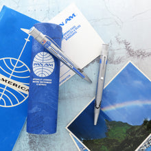 Load image into Gallery viewer, Two rollerball pens, one sits atop its presentation tube and the other one on top of the background, made of postcards and a map.