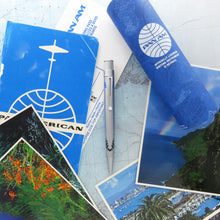 Load image into Gallery viewer, One rollerball pen displayed on top of the background, made of postcards and a map, the presentation tube is on the right side corner of the image.