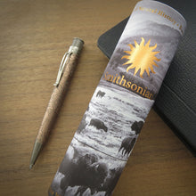 Load image into Gallery viewer, The Smithsonian National Museum of Natural History presentatnion tube lying on a wooden surface next to the rollerball pen, with a black leather-like object behind it. The cover shows bison imagery and the word Smithsonian.