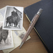 Load image into Gallery viewer, The acid-etched bison pen with a brushed brass clip and tip lies across a black leather notebook, placed on a wooden surface with two black-and-white photo prints nearby, and a vintage postcard.