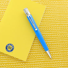 Load image into Gallery viewer, A blue retractable pen with chrome accents lies diagonally across a bright yellow notebook, which is placed on a yellow background with a repeating white wave pattern.