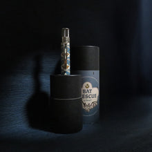 Load image into Gallery viewer, The pen placed inside the bottom half of the gift tube, which serves as a stand. The cap is set next to it on a low lit blue-colored background.