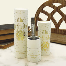 Load image into Gallery viewer, The pen in its presentation tube decorated with gold foil text reading 'The Cathedral' and a circular gold emblem, arranged on a marble surface with books and a wooden screen in the background.