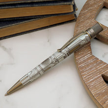 Load image into Gallery viewer, An ornate pen with intricate engravings, resting on a white marble surface next to a wooden object and a stack of vintage books.