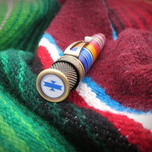 Load image into Gallery viewer, A close-up of the pen with a brushed brass cap featuring an Aztec-style graphic on top disc, resting on a textured knit blanket with red, green, and blue stripes.