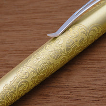 Load image into Gallery viewer, A close-up of the gold color pen with an embossed floral pattern, resting diagonally on a brown wooden surface.
