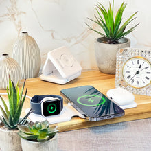 Load image into Gallery viewer, A wireless charging station with a smartphone, earbuds, and a smartwatch being charged simultaneously. The station is placed on a wooden surface with a marble background and decorative plants around.