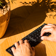 Load image into Gallery viewer, The keyboard extended on a wooden table next to a potted plant. A pair of hands are typing on the keys.