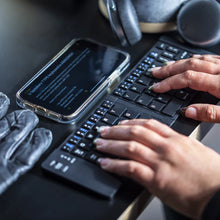 Load image into Gallery viewer, The keyboard is extended on a black table behind a cellphone. A pair of hands type on the keys.