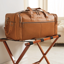 Load image into Gallery viewer, Angled view of the tan leather duffle bag set on a foldable wooden stand in a hotel setting.