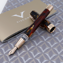 Load image into Gallery viewer, The Fountain pen set resting on top of the cap. Both are placed atop the presentation box and the included booklet. The Pen's barrel is made of light and dark red brown lacquer swirls with golden colored accents.