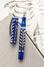 Load image into Gallery viewer, Front view of the Visconti Skeleton Sapphire Blue Fountain Pen with the cap set to the side. The background has the faded print of a skeleton's ribs.