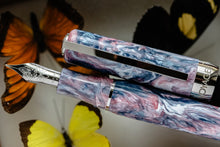 Load image into Gallery viewer, Close up to the grip section of the Visconti Voyager Mariposa Collection Fountain pen. It has silver colored accents, and the barrel is made of lacquer in pink, white, and blue swirls. Set on a background with butterflies.