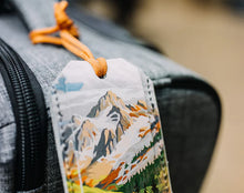Load image into Gallery viewer, Backpack with a mountain-themed tag on a blurred background