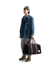 Load image into Gallery viewer, A young man holding the black duffle bag by the grab handles on his left hand.