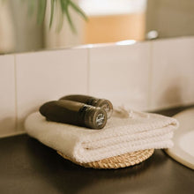 Load image into Gallery viewer, Two black travel bottles on top of a folded towel in a bathroom setting.
