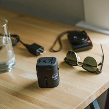 Load image into Gallery viewer, The black power adapter arranged on a wooden table with a camera, a charger, a water glass and some sunglasses.