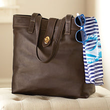 Load image into Gallery viewer, Angled front image of the brown leather tote bag. A pair of sun glasses and a blue striped scarf are hanging from the corner.