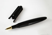 Load image into Gallery viewer, The rollerball with the cap set behind it on a white background. It is a thin oval shaped black pen. The nose is golden, and the cap has three different clips which can be used to stand it straight.