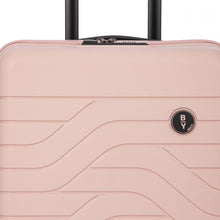 Load image into Gallery viewer, Close up to the top front of the pink pearl hard shell suitcase. The wave lines design is visible with the circular black BY logo at the top right.