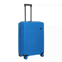 Load image into Gallery viewer, Front angled view of the electric blue hard shell suitcase with black trims. The panel has a textured wave lines design, and a small circular black logo with the letters BY on it. The telescopic handle is pulled up.