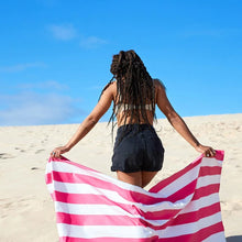 Load image into Gallery viewer, A young woman walking on sand dunes while carrying the white and pink stripes towel behind her.