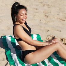 Load image into Gallery viewer, A young smiling woman wearing a black swimming suit sits at the beach on top of the green and white stripes towel.