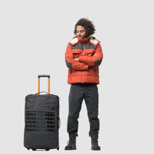 Load image into Gallery viewer, A man wearing an orange jacket standing next to the suitcase on a light gray background.