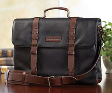 Load image into Gallery viewer, A black leather briefcase with brown accents, featuring a shoulder strap and a flap closure on a tabletop.