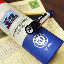 Load image into Gallery viewer, Top view of the pen leaning on the gift tube with a set of postcards arranged under. The top disk has the US Mail Eagle logo in it.