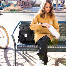 Load image into Gallery viewer, A young woman sitting on a public bench with the black backpack placed next to her.