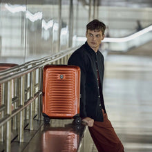 Load image into Gallery viewer, A young man leaning against a metallic railing with an orange suitcase.