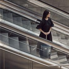 Load image into Gallery viewer, A young woman wearing a black dress heading down on an escalator with her navy blue suitcase.